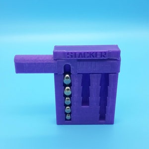 May include: Purple plastic toy with the word "STACKER" printed on the top. The toy has a sliding mechanism and five silver balls inside.