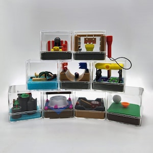 May include: A set of nine clear plastic cubes, each containing a miniature maze with a small ball. The mazes feature various themes, including a beach, a castle, a golf course, and a skate park.