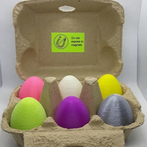 Puede incluir: Una huevera de cartón abierta con seis objetos coloridos en forma de huevo. Los huevos son de color rosa, verde, blanco, morado, amarillo y gris. Una etiqueta verde con texto es visible en la huevera. El texto de la etiqueta dice "No exponer a imanes."