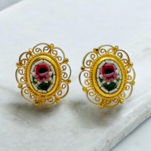 May include: A pair of gold earrings with a floral design. The earrings are made of filigree and have a small, oval-shaped center piece with a pink and green mosaic flower.