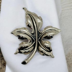 May include: A silver-toned leaf-shaped brooch with intricate details. The brooch is made of metal and has a smooth, polished finish.