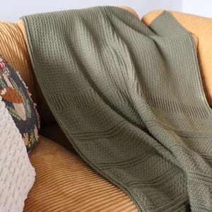 Könnte beinhalten: Eine grüne gestrickte Wolldecke, die über ein senfgelbes Cord-Sofa mit zwei dekorativen Kissen gelegt ist.