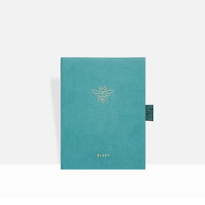 Puede incluir: Diario de color turquesa con un emblema de abeja dorada y la palabra "DIARY" impresa en la parte inferior. El diario tiene un cierre de tela turquesa a juego. La cubierta tiene una textura suave y aterciopelada.