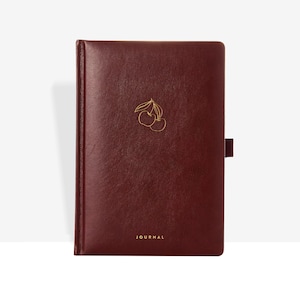 Puede incluir: Un diario burdeos con una ilustración de cerezas doradas y la palabra "JOURNAL" en letras doradas. El diario tiene una textura similar al cuero y un cierre magnético.