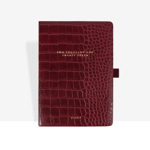 Könnte beinhalten: Ein tiefburgunderrotes Tagebuch mit einer strukturierten Krokodilleder-Optik auf dem Einband. Auf der Vorderseite befindet sich der goldene Schriftzug TWO THOUSAND AND TWENTY SEVEN und DIARY. Eine kleine Stiftschlaufe aus Stoff befindet sich rechts.