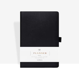 May include: A black planner with a white band featuring the word "PLANNER" in gold lettering. The planner has a black elastic band closure and the text "NO DATES - START ANYTIME" is also visible.