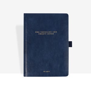 May include: A dark blue diary with a textured cover featuring the gold embossed text TWO THOUSAND AND TWENTY SEVEN at the top and the word DIARY at the bottom. The notebook includes a dark blue elastic pen loop attached to the right side edge.