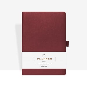 Può includere: Un'agenda bordeaux con una fascia bianca con la scritta "PLANNER" e il testo "NO DATES - START ANYTIME". L'agenda ha una chiusura elastica bordeaux abbinata. L'agenda è di Scribble &.