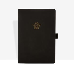 Op de afbeelding: Een zwart notitieboek met een gouden bijenlogo op de kaft. Het notitieboek heeft een zwarte elastische sluiting. De kaft is glad en het bijenlogo is een eenvoudige lijntekening.