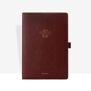 Può includere: Un diario in pelle bordeaux con un'emblema a forma di ape dorata. La parola "DIARY" è stampata in oro in basso. Il diario ha una chiusura magnetica sul lato destro.