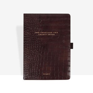 May include: A dark brown diary with a textured crocodile skin pattern. The cover features gold foil text that reads TWO THOUSAND AND TWENTY SEVEN at the top and DIARY at the bottom. A small fabric pen loop is attached to the right side of the cover.