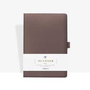 Op de afbeelding: Een gesloten, rechthoekige planner met een bruine kaft en een witte band in het midden. De band bevat het woord "PLANNER" en de tekst "50 DATES - START ANYTIME". Een bruine stoffen bladwijzer is aan de rechterkant bevestigd.