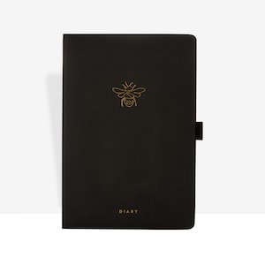 May include: Black diary with a gold bee emblem and the word "DIARY" at the bottom. The diary has a black strap closure on the right side. The bee emblem is centered at the top of the diary.