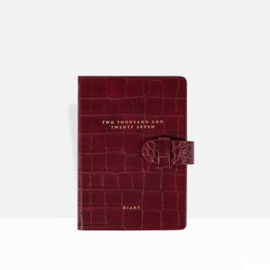 May include: A deep burgundy diary with a textured crocodile-embossed finish. The front cover features the gold-stamped text "TWO THOUSAND AND TWENTY SEVEN" and "DIARY" at the bottom. A matching strap with a snap closure secures the front cover.