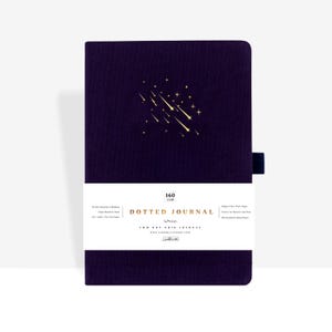Pode incluir: Um diário pontilhado roxo escuro com um design de estrela cadente dourada. O diário tem uma faixa branca com o texto "DOTTED JOURNAL" e outros detalhes. Uma fita de marcador azul escuro está anexada.