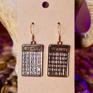 May include: Gold-colored calculator earrings with the number 3.14159 displayed on the top. The earrings are rectangular and hang from gold-colored hooks. The earrings are displayed on a tan card.