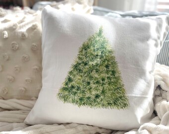May include: A white throw pillow with a green watercolor Christmas tree design. The pillow is on a white fluffy blanket.