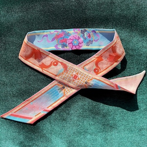 May include: A long, narrow silk scarf featuring a vibrant design with orange dragon motifs, geometric patterns, and light blue sections decorated with pink floral elements and small insect illustrations. The fabric has a smooth, satin-like finish.