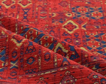 May include: Close-up of a red fabric with a detailed geometric pattern. The design incorporates blue and gold shapes, creating a textured appearance. The fabric is folded, showing the intricate weave and color variations.