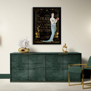 May include: A framed art print featuring a woman in a blue dress holding a red object, with the text "PLEASE DON'T DO COCAINE IN THE BATHROOM" in gold lettering. The artwork is displayed above a dark green cabinet.