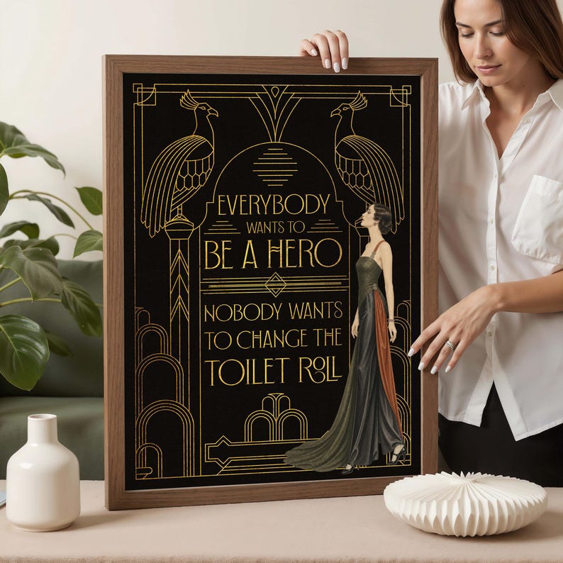 Pu&ograve; includere: Un quadro rettangolare incorniciato con sfondo nero, linee geometriche dorate in stile Art D&eacute;co e due pavoni. Il testo recita: EVERYBODY WANTS TO BE A HERO NOBODY WANTS TO CHANGE THE TOILET ROLL. &Egrave; raffigurata una donna che indossa un abito lungo.