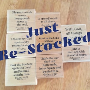 May include: A collection of eight square wooden rubber stamps, each featuring a different religious quote and scripture reference printed in black ink. The stamps are arranged in a cluster, showcasing light-colored wood blocks with text.