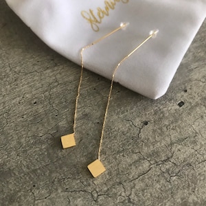 May include: A pair of gold threader earrings. Each earring has a fine chain extending from a post, with a small square pendant at the end. The earrings are presented on a white pouch with gold script.