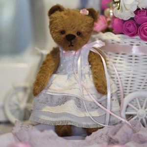 May include: A brown teddy bear wearing a white dress with pink lace trim and a pink ribbon. The bear is sitting in a white wicker basket with pink flowers.