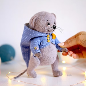 May include: A gray plush mouse wearing a blue and white polka dot hooded jacket with yellow buttons. The mouse is standing on a white surface with string lights.