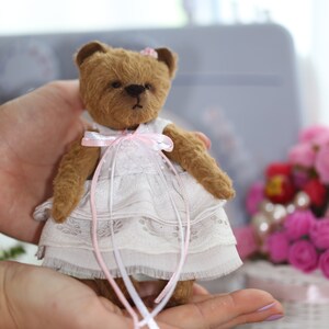 May include: A small brown teddy bear wearing a white dress with pink lace trim and a pink ribbon around its neck. The bear is being held in someone's hands.