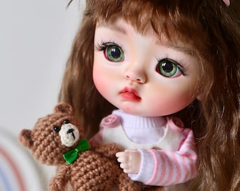 May include: A ball-jointed doll with long wavy brown hair, green eyes, and long eyelashes, wearing a pink and white striped long-sleeve shirt under a cream-coloured pinafore dress with a bear graphic, holding a small brown crocheted teddy bear.