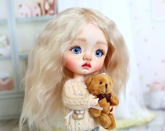 May include: A handmade doll with long, wavy blonde hair and striking blue eyes. The doll is wearing a beige sweater and holding a small brown teddy bear with a dark brown bow. The doll has rosy cheeks and a delicate expression.