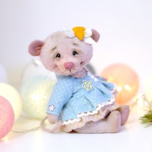 May include: A small, pink plush mouse wearing a blue dress with white polka dots and a yellow star. The mouse has a white bow with a yellow flower on its head. The mouse is sitting on a white surface with a string of lights behind it.