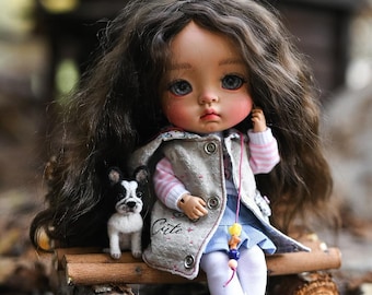 May include: A doll with long brown hair and blue eyes, wearing a gray vest, pink and white striped shirt, and blue skirt. A small Boston Terrier figurine sits beside the doll. The vest has the word "Cute" written on it. The doll is sitting on a wooden bench.