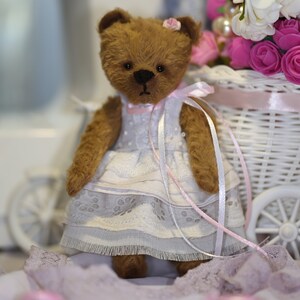 May include: A brown teddy bear wearing a white dress with pink lace trim and a pink ribbon. The bear is sitting in a white wicker basket with pink flowers.