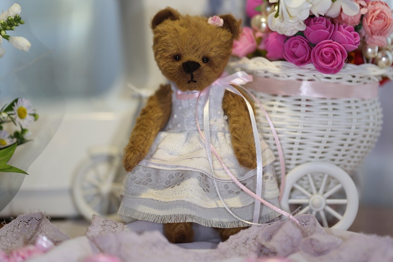 May include: A brown teddy bear wearing a white dress with pink lace trim and a pink ribbon. The bear is sitting in a white wicker basket with pink flowers.
