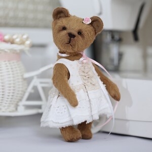 May include: A brown teddy bear wearing a white dress with lace trim and a pink flower on its head. The bear is standing on a white surface.