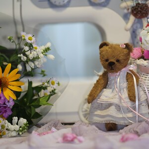 May include: A brown teddy bear wearing a white dress with pink accents sits in front of a white wicker basket filled with pink and white flowers. The bear is holding a pink ribbon in its paws.