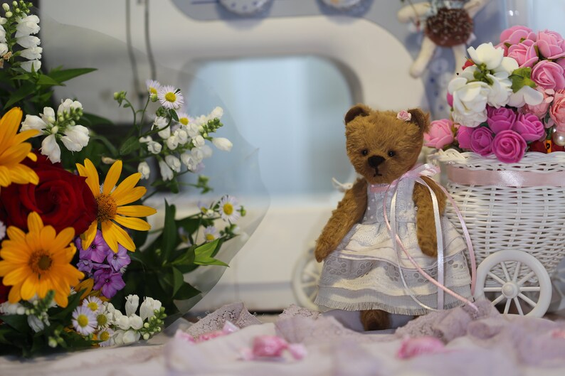 May include: A brown teddy bear wearing a white dress with pink accents sits in front of a white wicker basket filled with pink and white flowers. The bear is holding a pink ribbon in its paws.