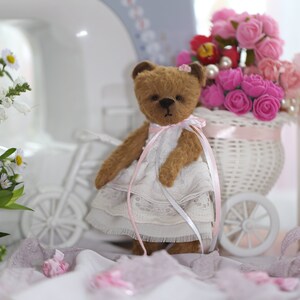 May include: A brown teddy bear wearing a white dress with pink lace trim and a pink bow. The bear is sitting in a white wicker basket filled with pink roses.