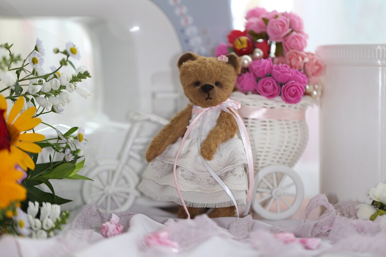May include: A brown teddy bear wearing a white dress with pink lace trim and a pink bow. The bear is sitting in a white wicker basket filled with pink roses.