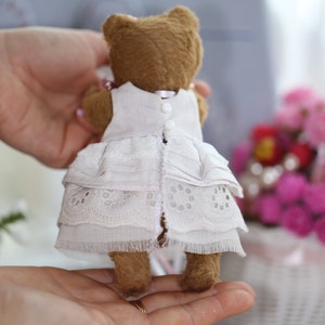May include: A brown teddy bear wearing a white lace dress with ruffles. The bear is being held by a person's hands.