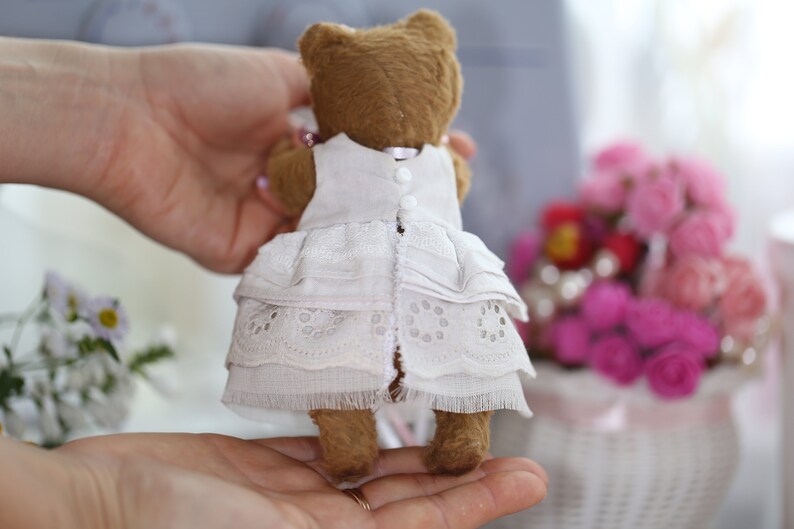 May include: A brown teddy bear wearing a white lace dress with ruffles. The bear is being held by a person's hands.