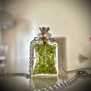 May include: A silver chain necklace with a clear glass bottle pendant filled with green peridot stones. The bottle has a silver crown-shaped stopper with a clear rhinestone.