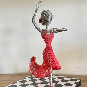May include: Wire sculpture of a dancer in a red dress, standing on a black and white checkered base.