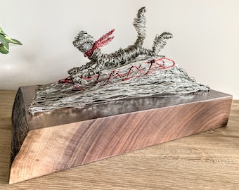 May include: A wire sculpture of a person sledding down a hill made of wire. The sculpture is on a wooden base.