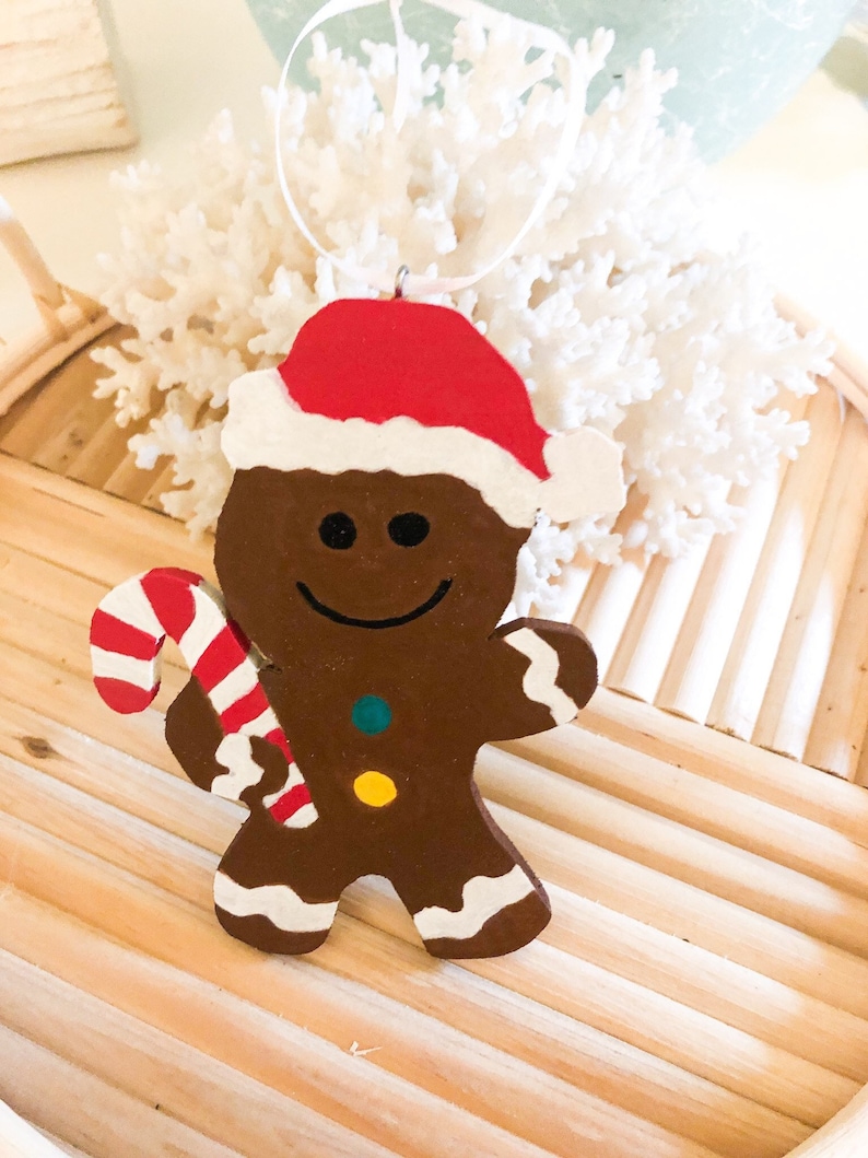 Puede incluir: Un adorno de hombre de jengibre marr&oacute;n con un gorro de Pap&aacute; Noel rojo y blanco. La figura tiene una cara sonriente negra, dos botones coloridos en el pecho y detalles de glaseado blanco en los pies y brazos. Sostiene un bast&oacute;n de caramelo.