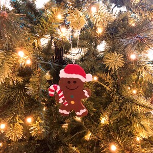 Puede incluir: Un adorno de hombre de jengibre marr&oacute;n con un gorro de Pap&aacute; Noel rojo y blanco, sosteniendo un bast&oacute;n de caramelo a rayas rojas y blancas. La figura presenta una cara sonriente, dos peque&ntilde;os botones de colores y detalles de glaseado blanco.