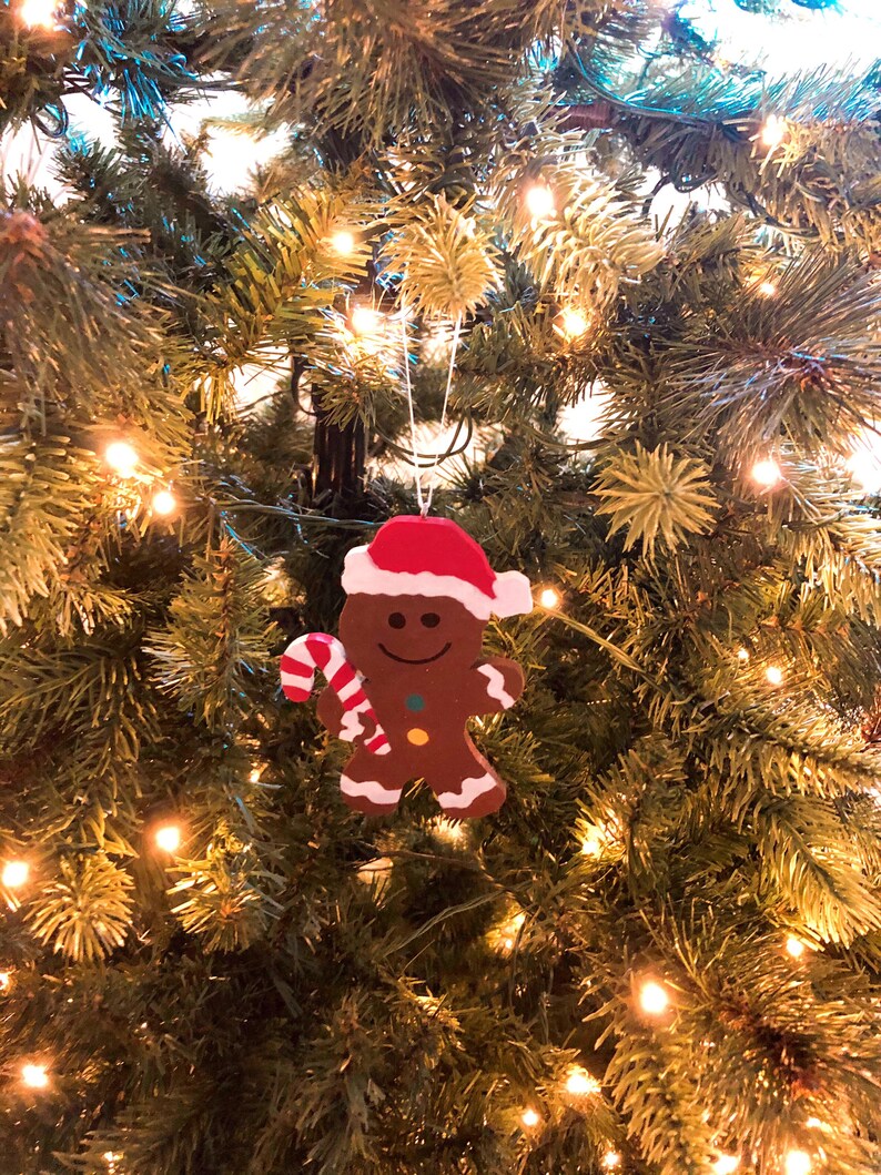 Puede incluir: Un adorno de hombre de jengibre marr&oacute;n con un gorro de Pap&aacute; Noel rojo y blanco, sosteniendo un bast&oacute;n de caramelo a rayas rojas y blancas. La figura presenta una cara sonriente, dos peque&ntilde;os botones de colores y detalles de glaseado blanco.