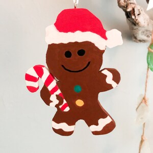 Puede incluir: Un adorno de hombre de jengibre marr&oacute;n con un gorro de Pap&aacute; Noel rojo y blanco. La figura tiene una cara sonriente, un bot&oacute;n verde y otro amarillo en el pecho, y detalles de glaseado blanco en los pies y brazos. Sostiene un bast&oacute;n de caramelo.